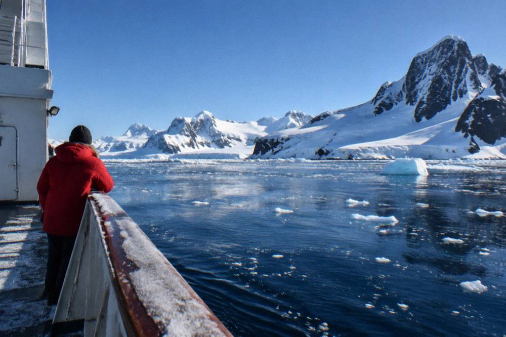 Person in a red parka standing at the edge of an expedition ship deck above icy water