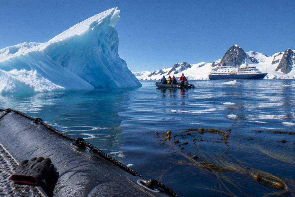 A Zodiac cruising near an iceberg with an expedition ship in the background under blue skies