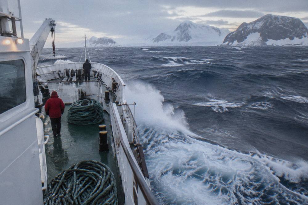 An expedition cruise ship moving through open ocean swells with grey skies above