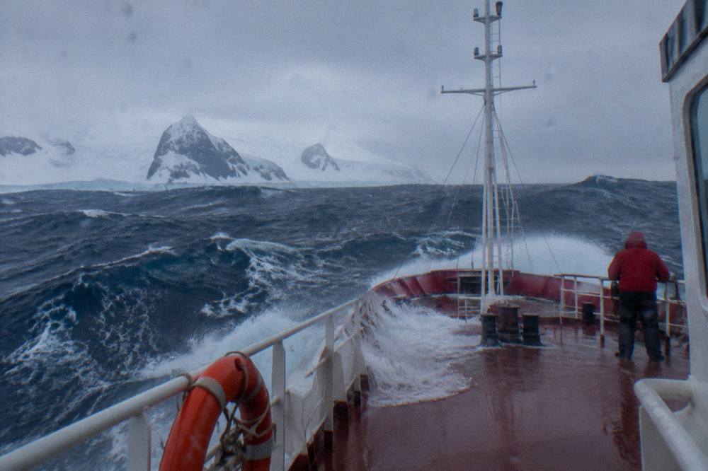 A small red-hulled expedition ship pushing through large grey waves in open water
