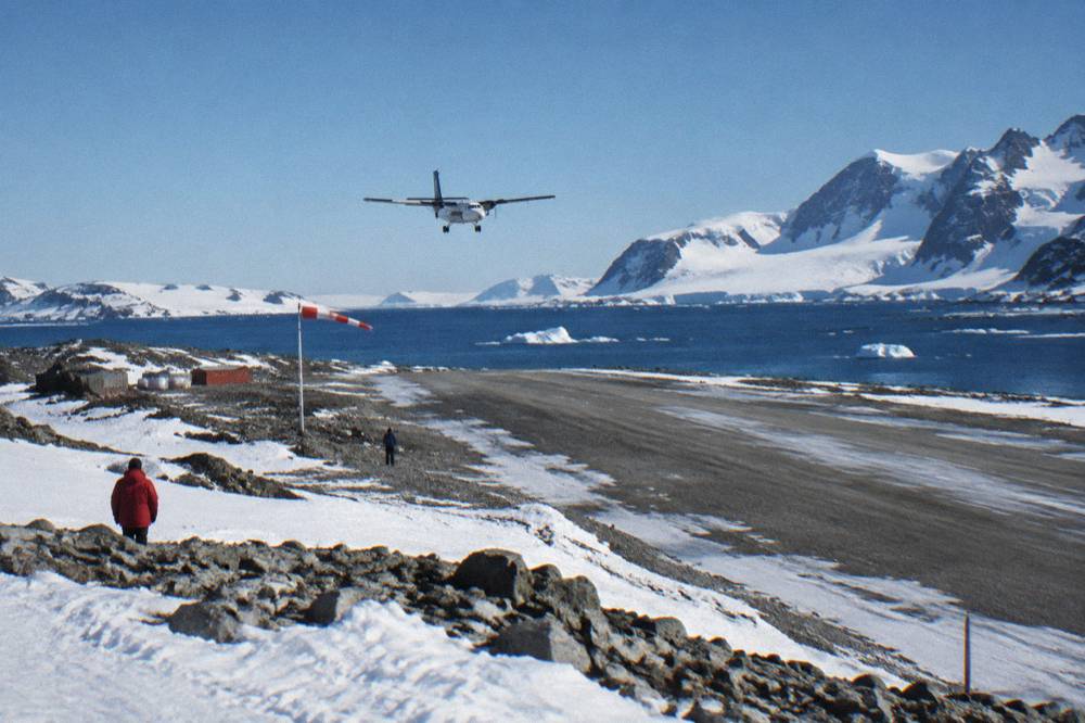 A twin-engine charter aircraft flying over cloud-covered ocean toward a snowy island airstrip