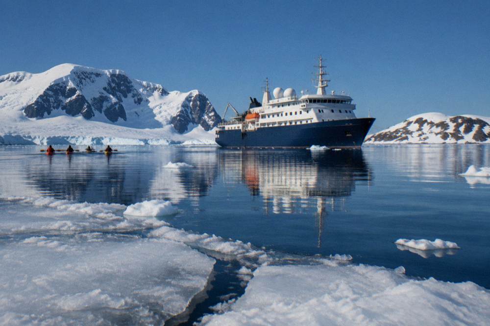 A polar-class expedition ship at anchor in a calm Antarctic bay surrounded by ice and low mountains
