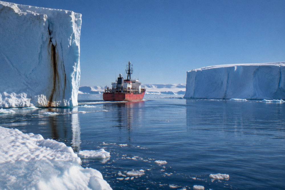 Small red expedition ship cruising between tall icebergs in calm polar water