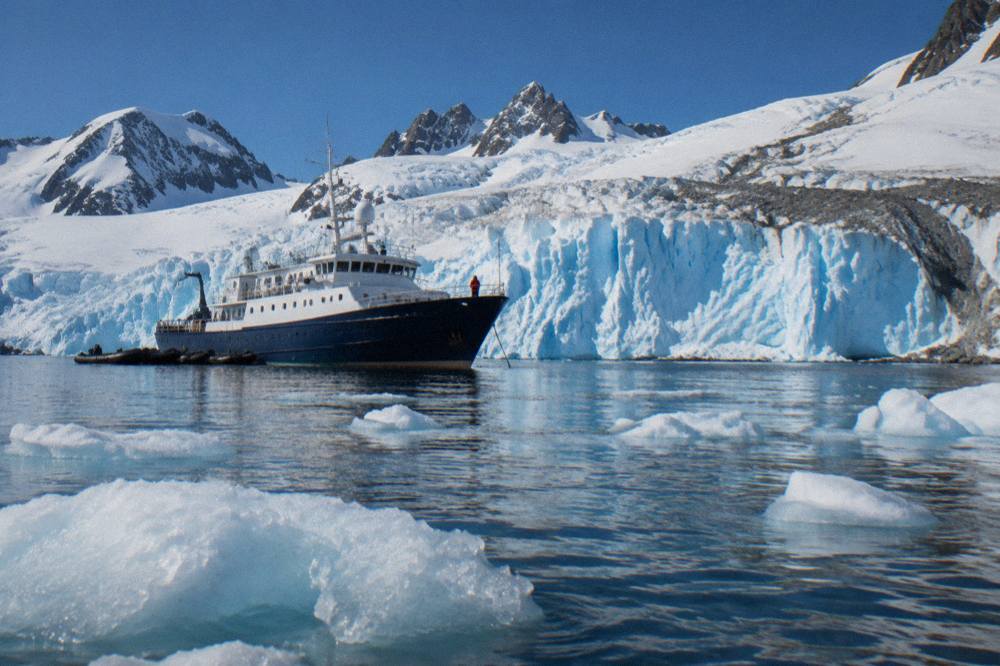 Small expedition ship at anchor near a glacier front with zodiacs parked alongside