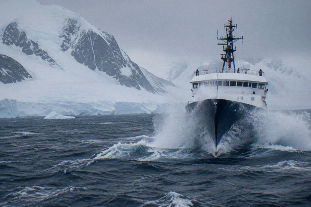 Expedition ship moving through open ocean swell with grey skies