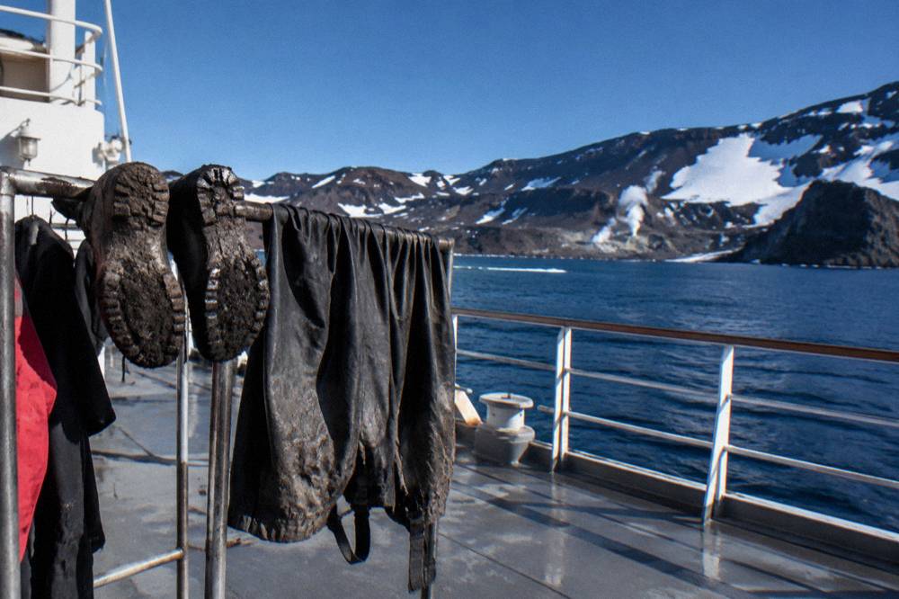 Muddy expedition boots and waterproof gear drying on a rack on a ship deck