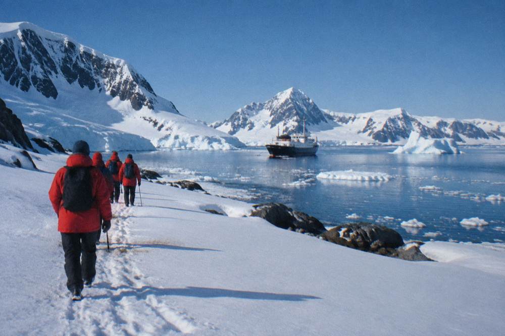 A group of travellers in red parkas walking along a snowy shoreline beside a small expedition ship