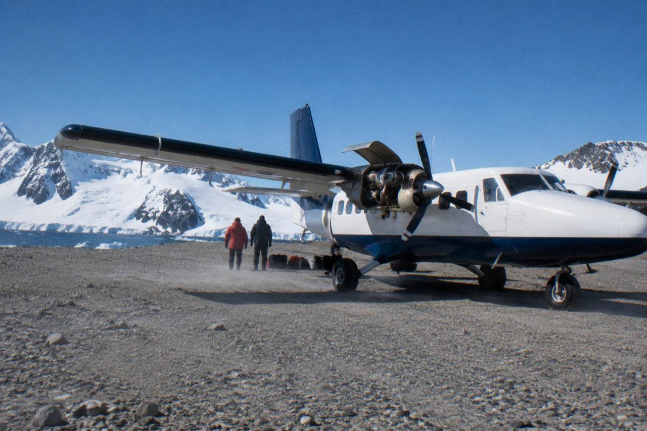 A small charter aircraft on a gravel runway on King George Island with snowy Antarctic mountains behind
