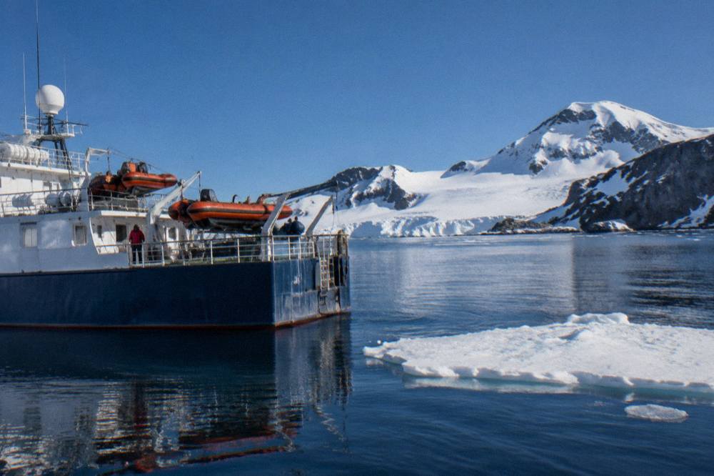 A small expedition ship at anchor in a calm Antarctic bay with Zodiacs ready at the marina deck.