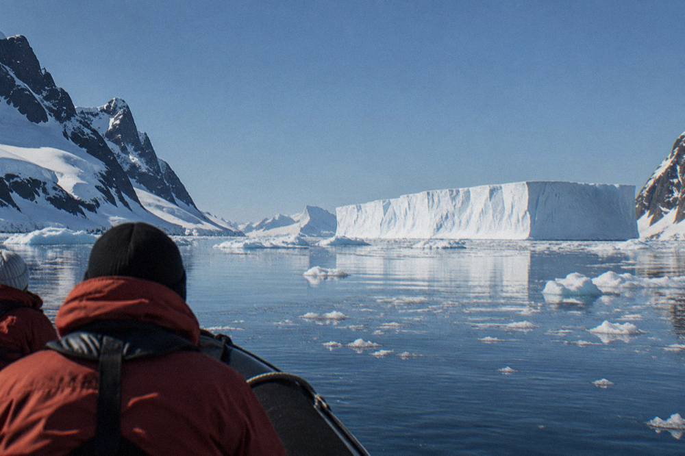 Flat calm polar water dotted with brash ice and a large tabular iceberg under a pale sky