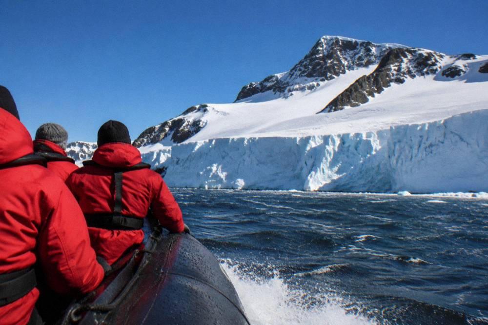 Zodiac cruising past a low ice cliff with expedition passengers in red parkas