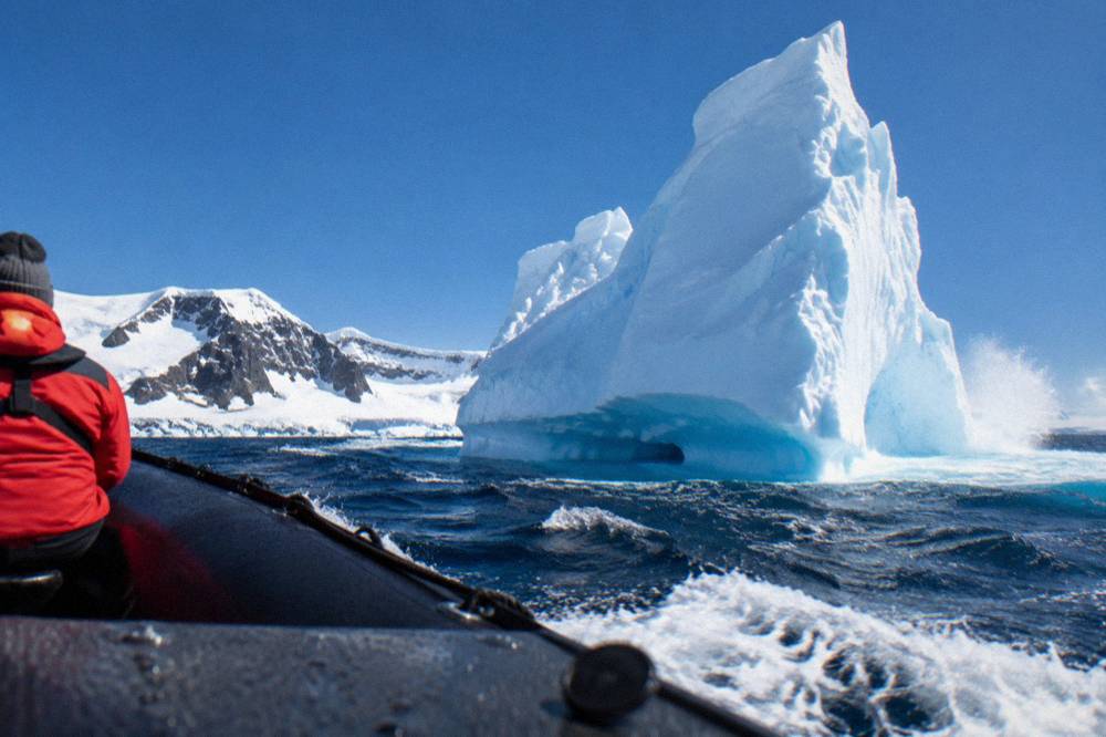 Zodiac cruising past a tall iceberg near the Antarctic Peninsula coastline