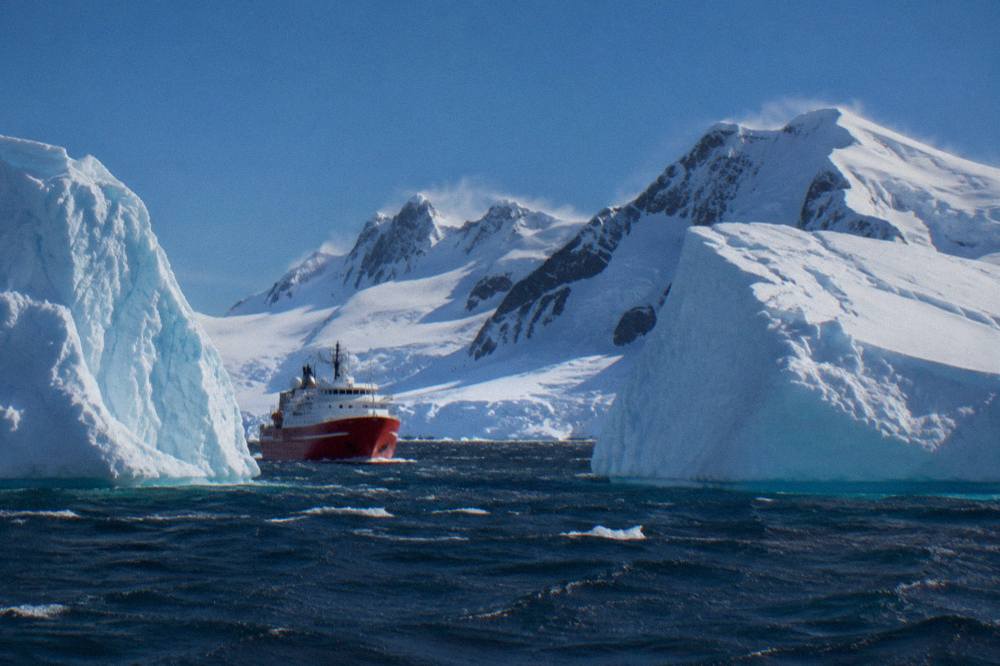 Small red-hulled expedition ship cruising past towering icebergs in calm polar water