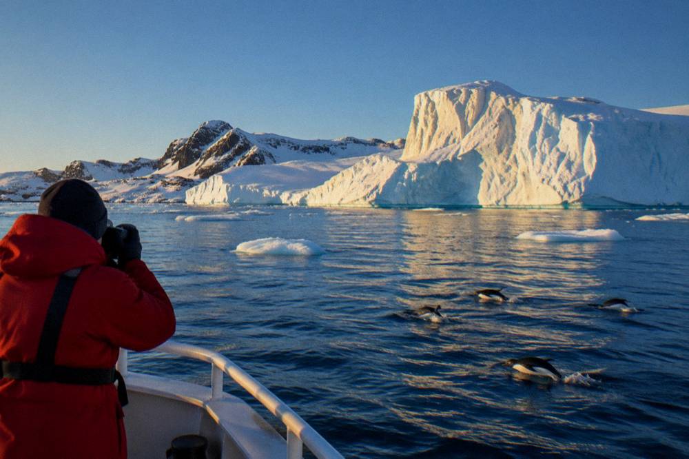 Traveller in expedition parka photographing an iceberg from the bow of a small ship