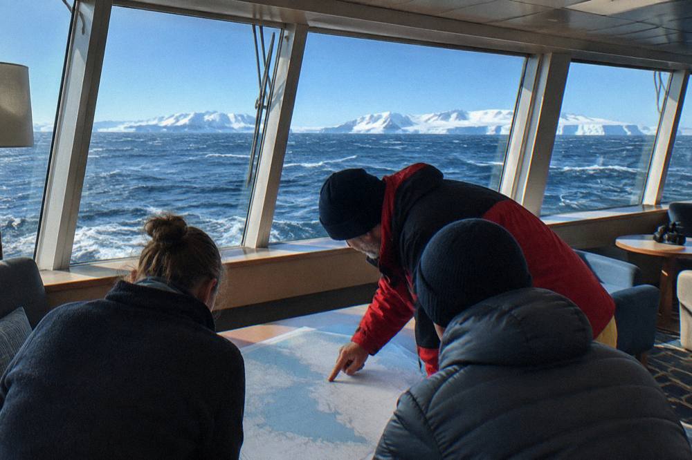 Couple reviewing a map with an expedition leader in a ship lounge with panoramic windows