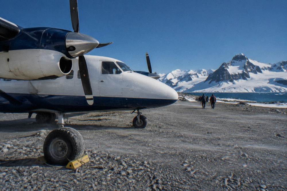 A small twin-engine aircraft on a gravel airstrip surrounded by snowy hills