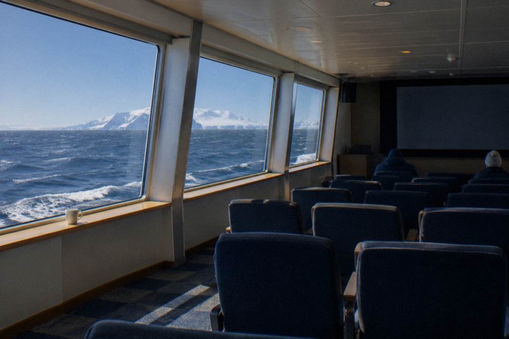 Empty expedition lecture lounge with rows of seats facing a screen and wide windows onto open polar sea