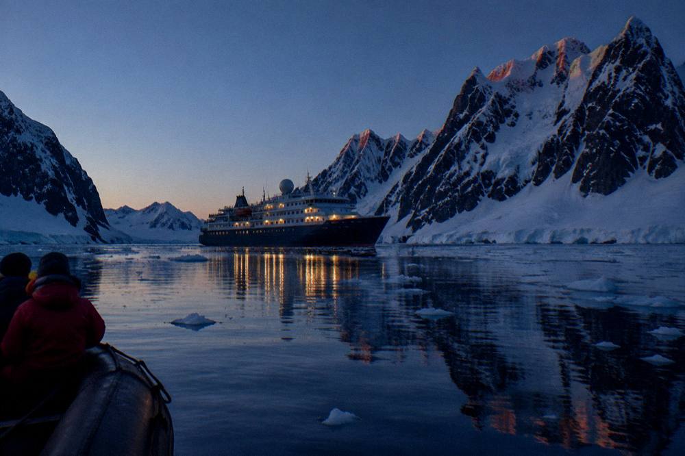 Expedition ship at anchor in a calm polar bay with jagged snow-covered peaks behind it