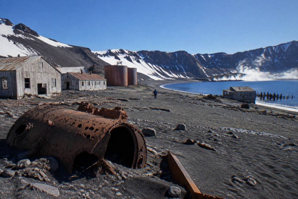 Abandoned wooden buildings and rusting tanks on a black volcanic beach inside an Antarctic caldera