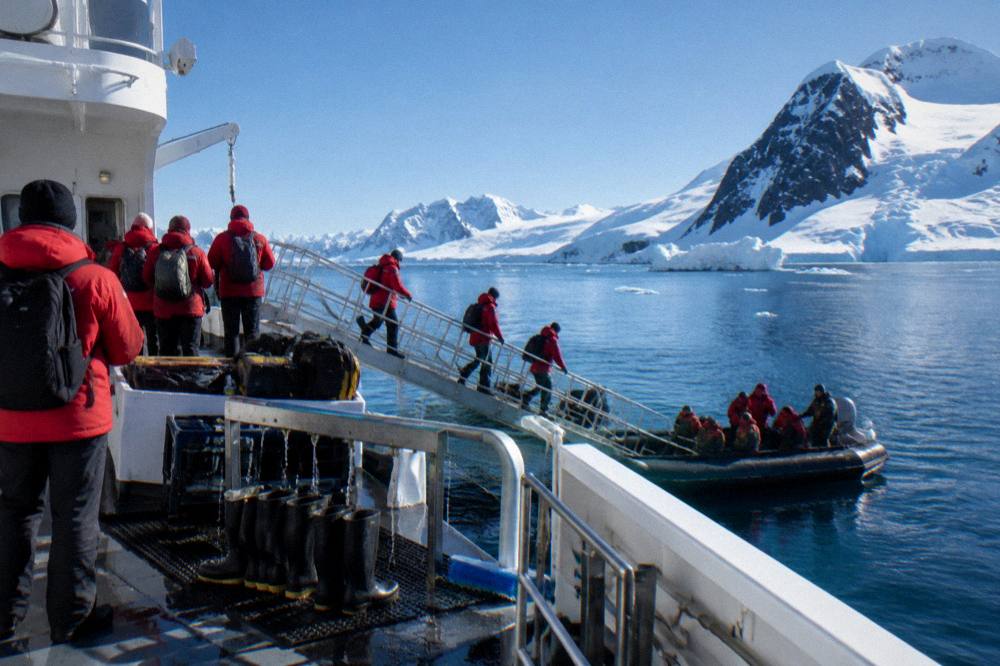 Expedition passengers in red parkas lined up at a ship gangway waiting to board Zodiacs