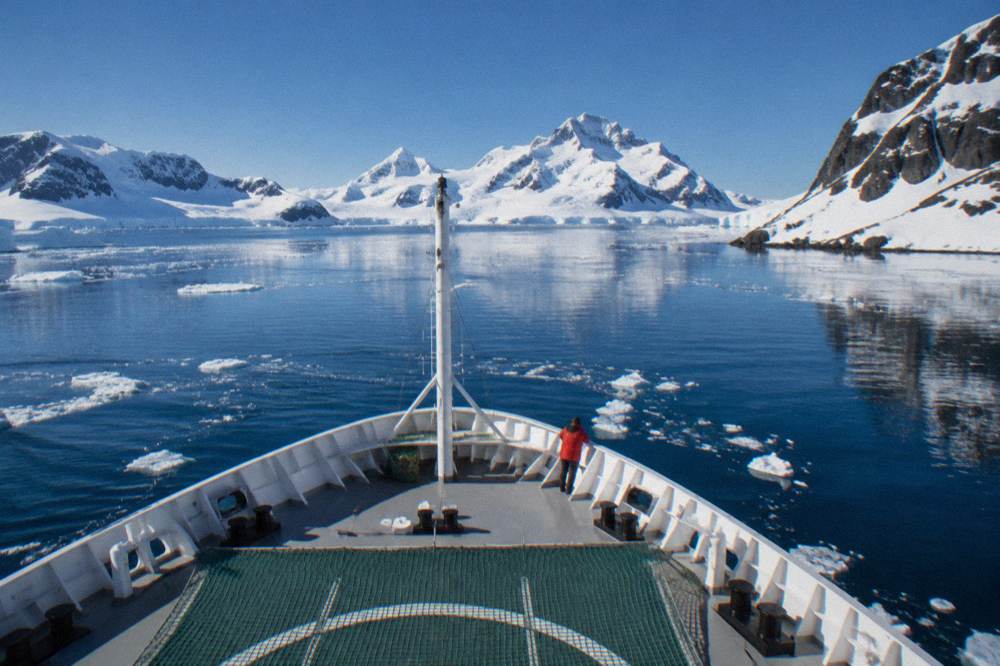 Expedition ship bow cutting through calm polar water with snow-covered peaks behind
