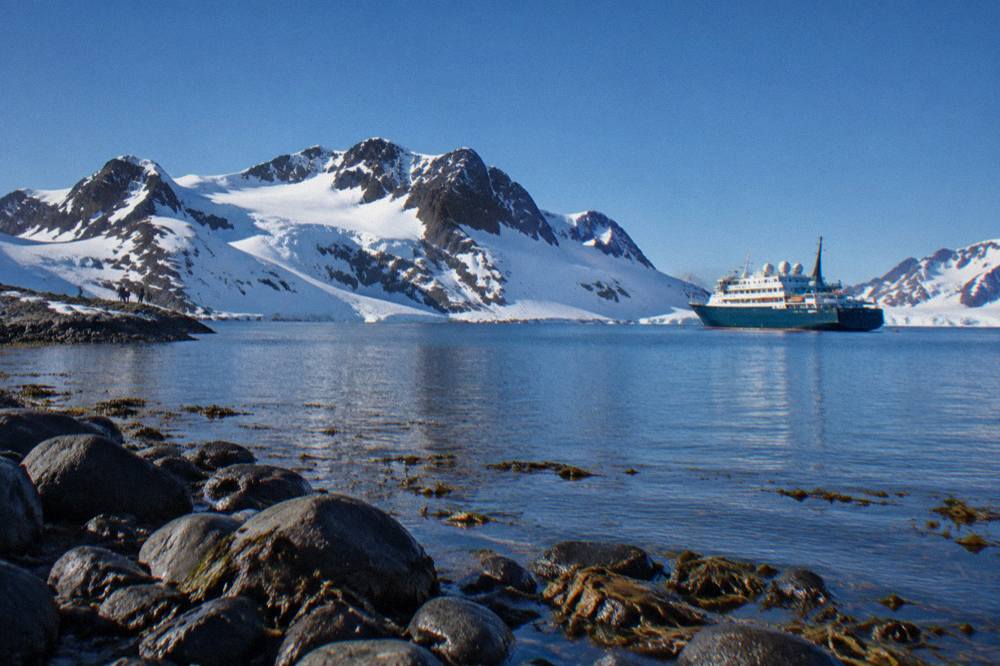 Small expedition ship at anchor in a calm Antarctic bay with ice-covered mountains behind it