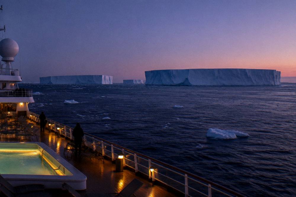 Elegant expedition ship with a lit pool deck cruising past tabular icebergs at dusk