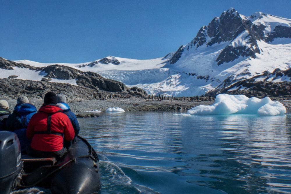Zodiac carrying visitors toward a rocky Antarctic shoreline with penguins in the distance