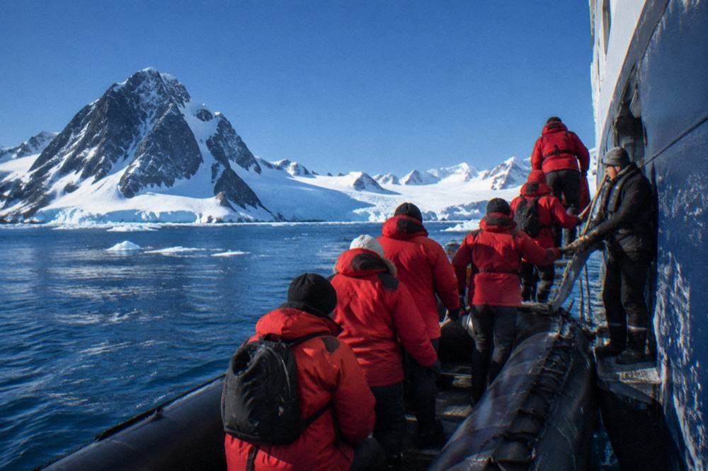 Expedition passengers in red parkas lining up at a Zodiac gangway beside an ice-covered ship