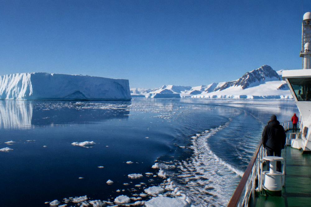 Small expedition ship moving through calm polar water between icebergs and mountains