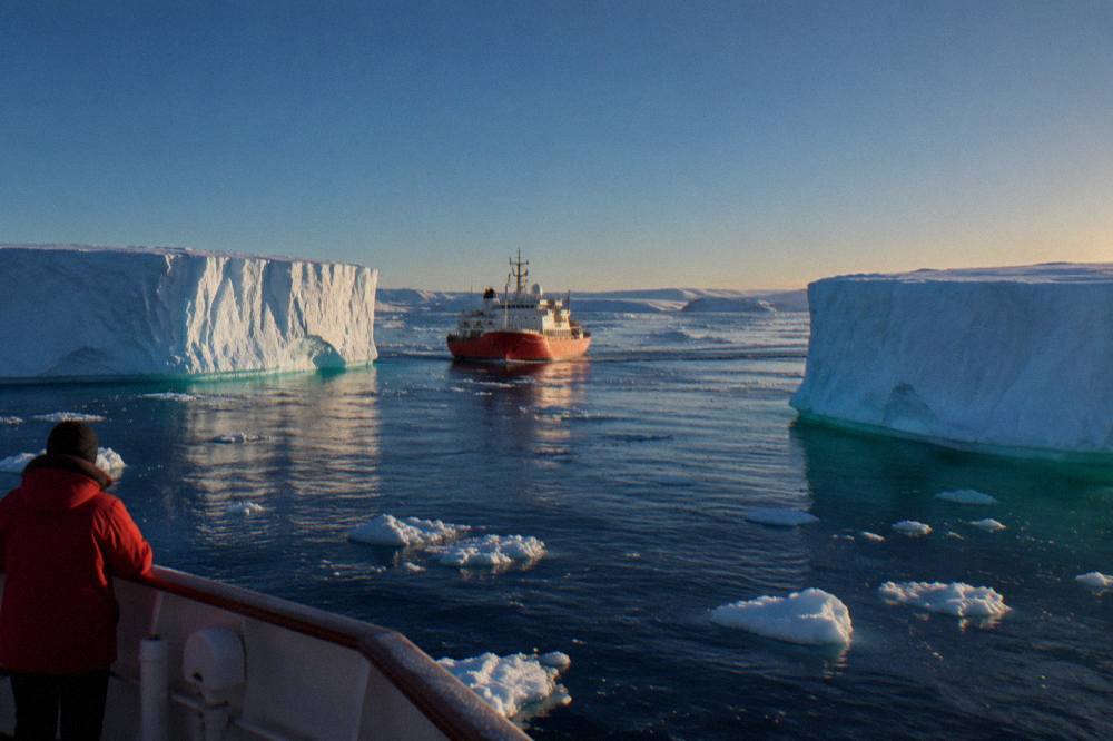 Small red-hulled expedition ship cruising past tabular icebergs in calm polar water