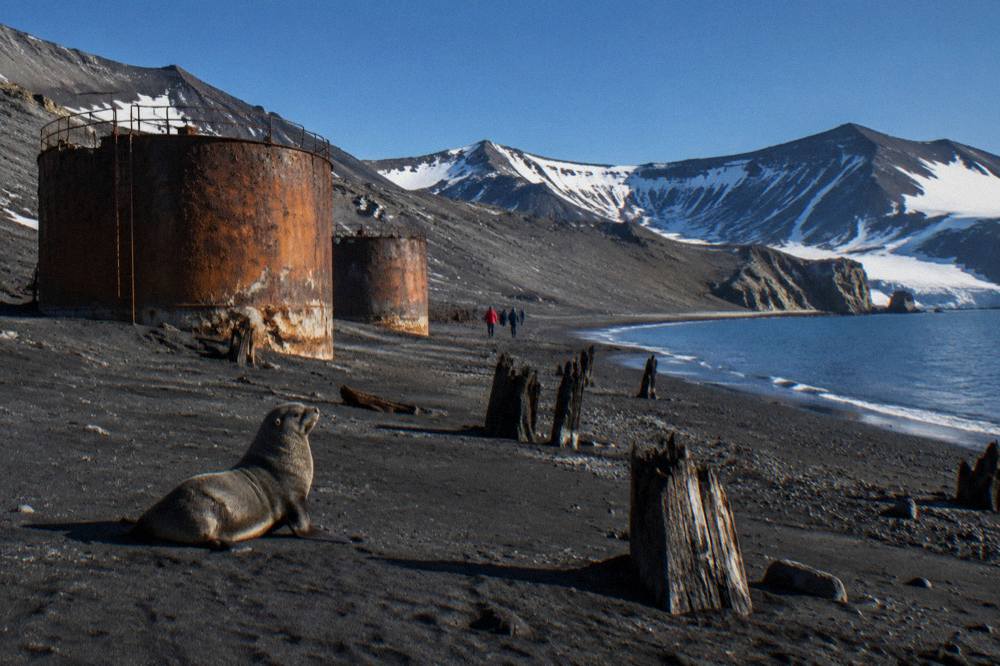 A dark volcanic beach with the rusted remains of wooden buildings and whale oil tanks along the shore