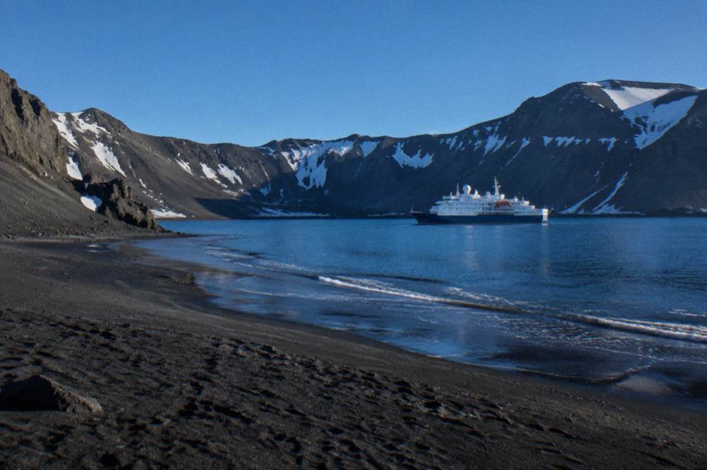 An expedition ship at anchor inside a circular volcanic bay with dark mountain ridges around the water
