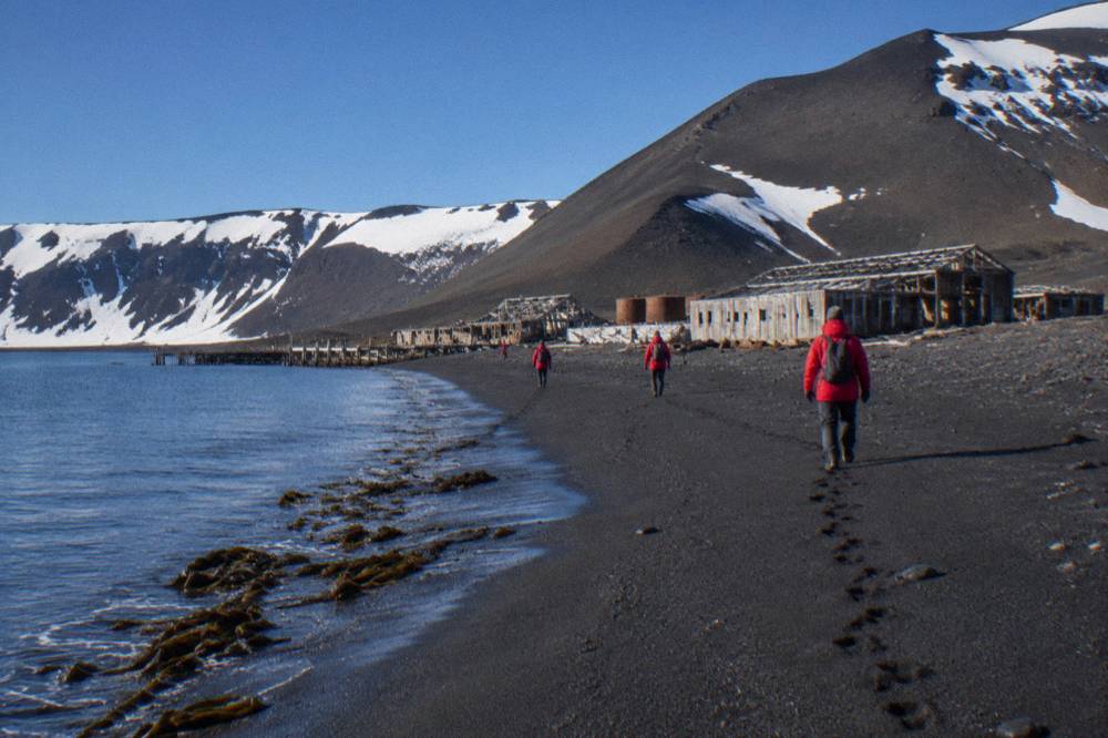 A group of passengers in red parkas walking along a black volcanic beach toward old wooden buildings