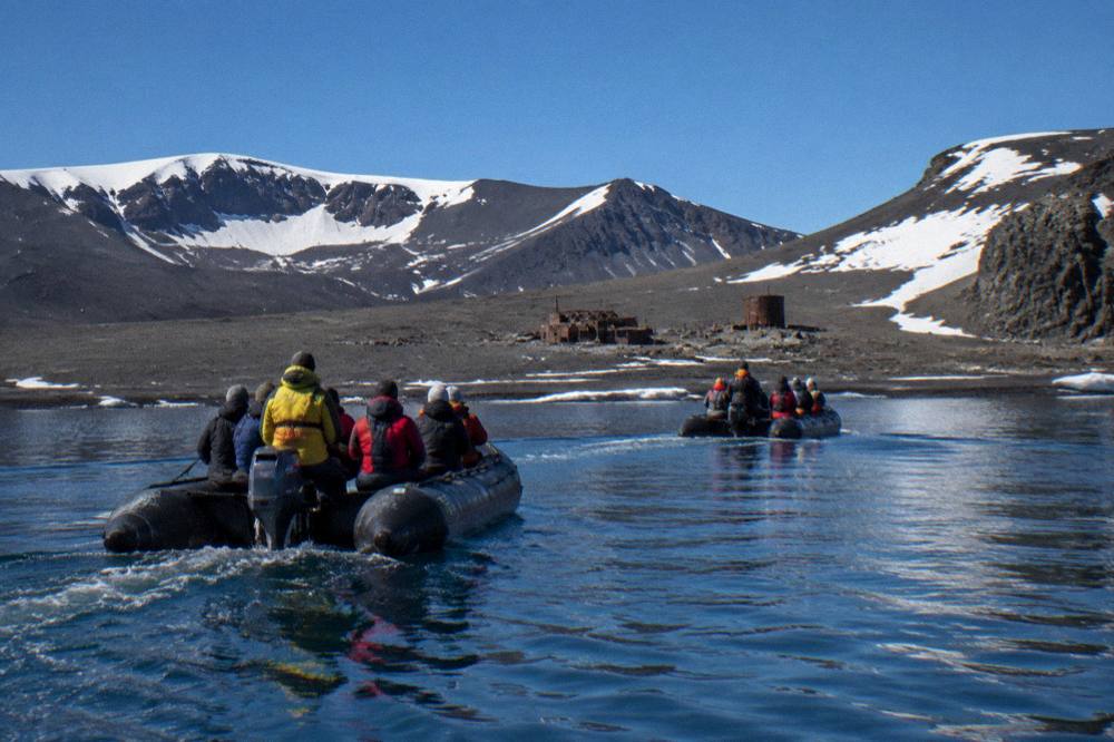 Zodiac boats carrying passengers across calm grey water toward a dark volcanic shoreline