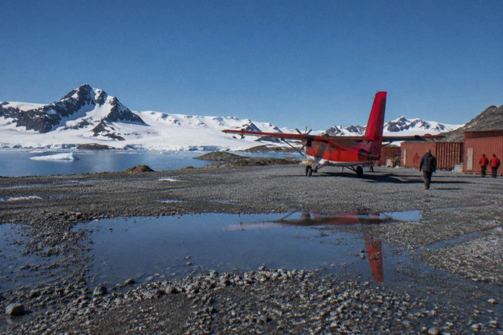 Small red charter plane on a gravel airstrip beside a polar ship at King George Island