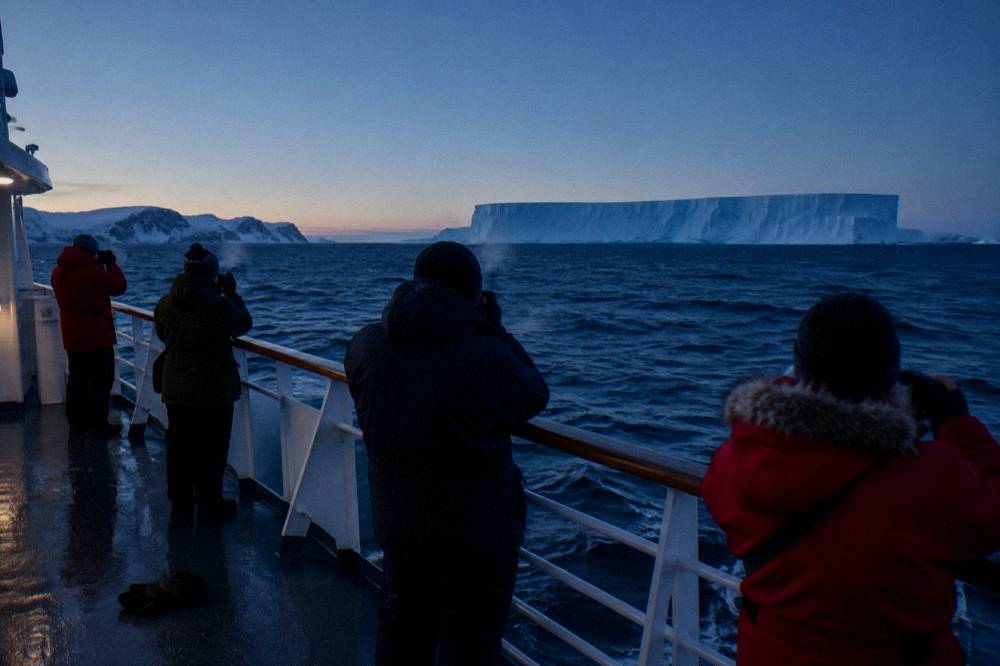 Passengers gathered at the ship rail photographing a large tabular iceberg at dusk