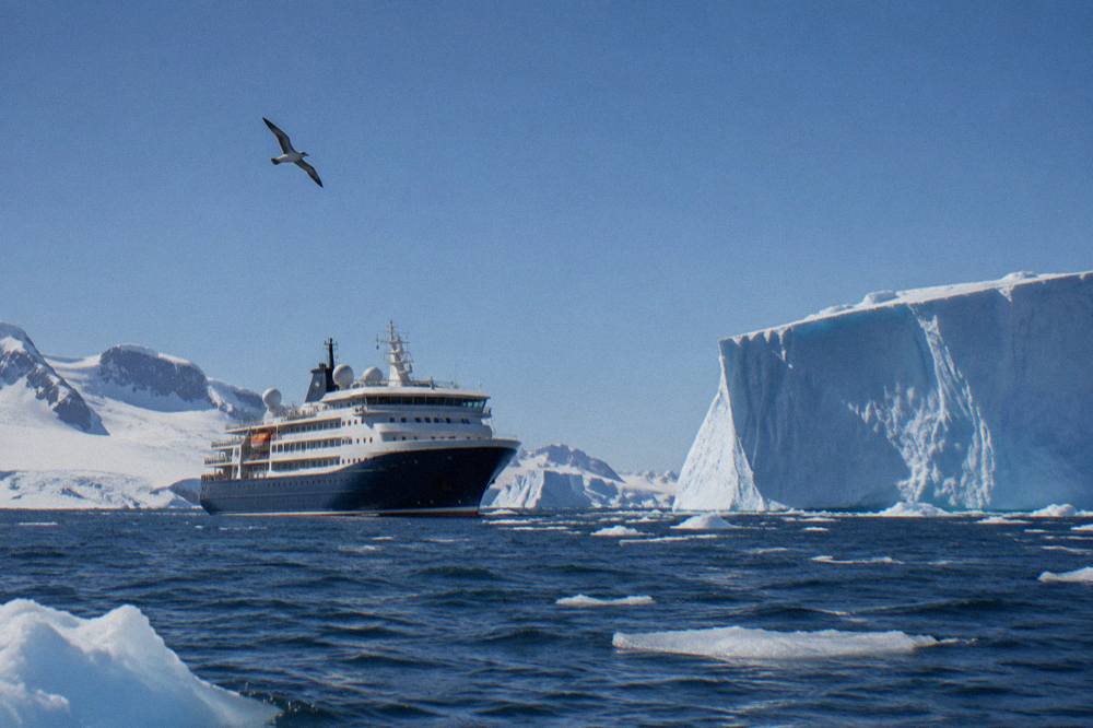 A modern expedition ship with a rounded bow cruising past tabular icebergs under a pale sky