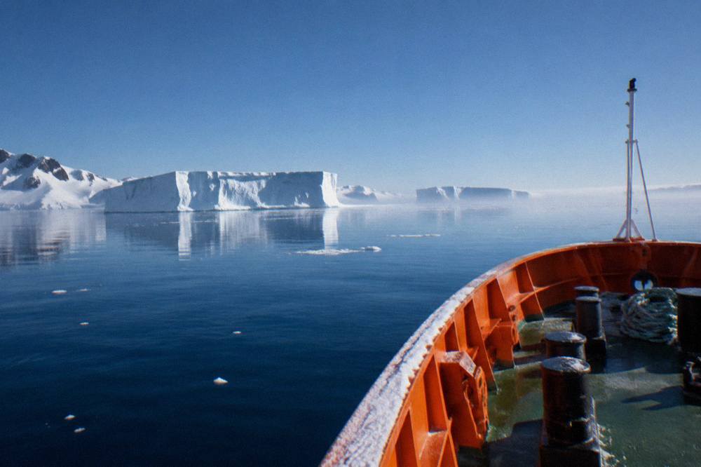 The bow of an orange expedition ship cutting through calm polar water with icebergs in the distance