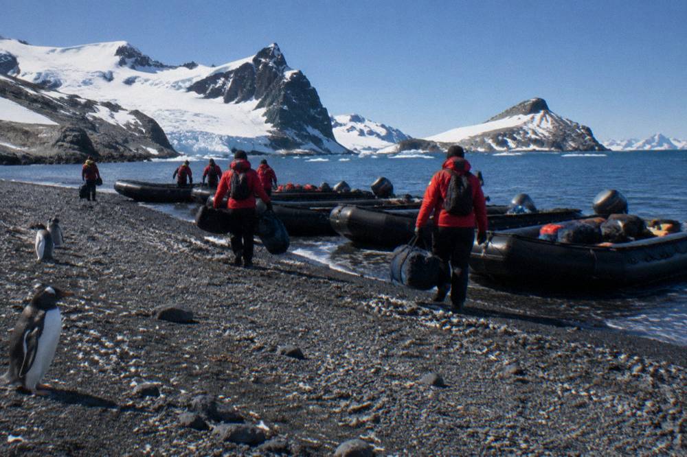 Zodiacs lined up on a rocky Antarctic beach with passengers in red parkas nearby
