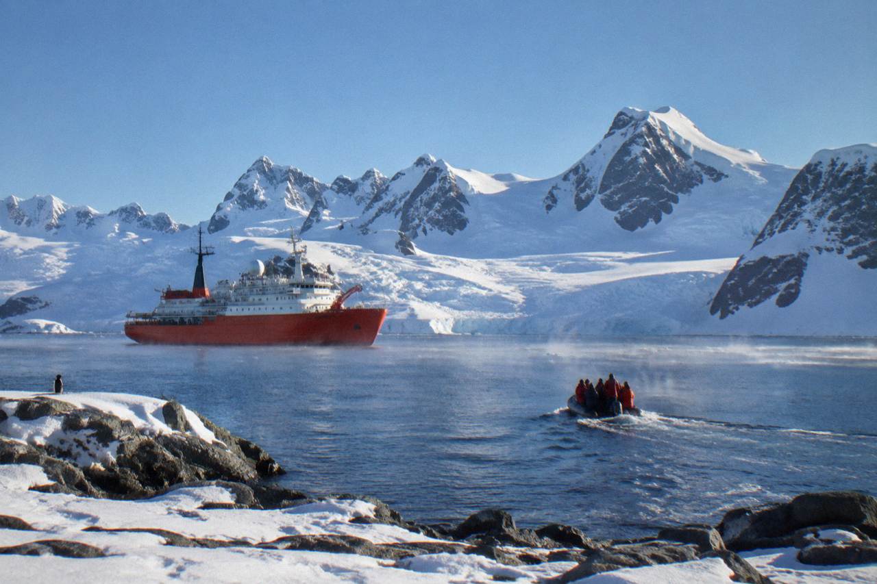 An orange-hulled expedition ship anchored near snow-covered Antarctic mountains