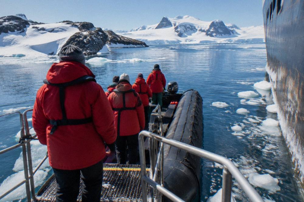 Passengers in red parkas boarding a Zodiac from an expedition ship gangway in calm polar water