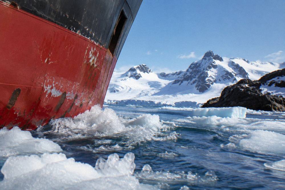 An expedition ship's bow cutting through calm icy water with snow-covered mountains beyond