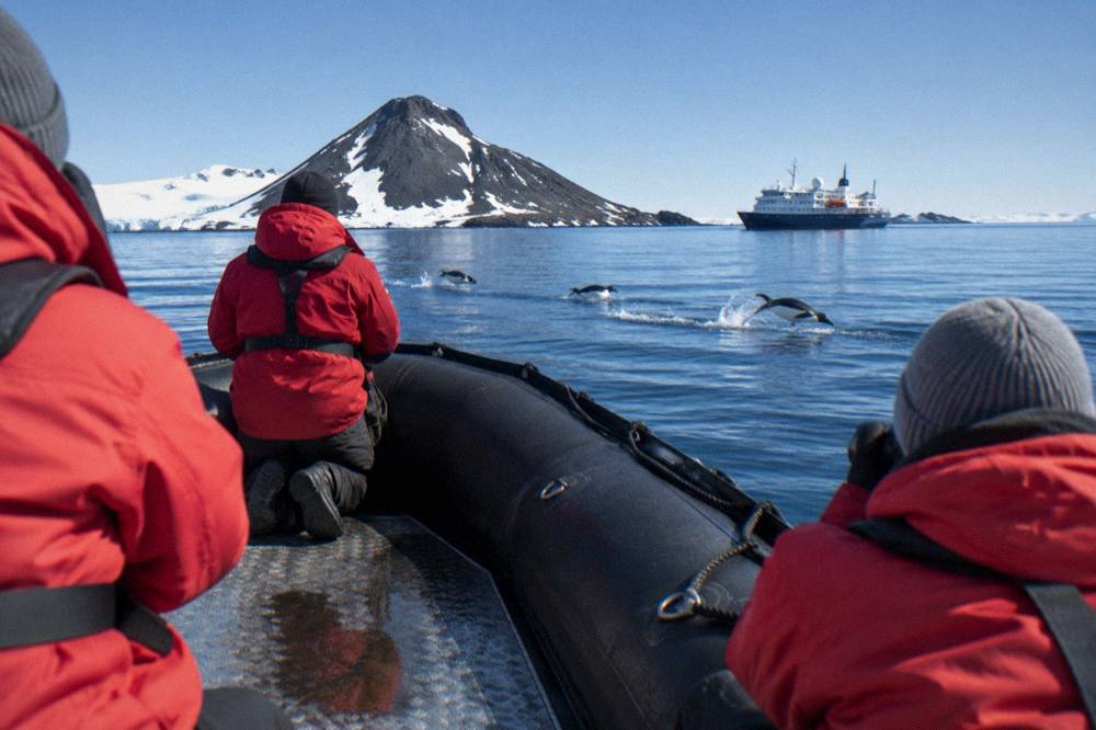 Passengers in red parkas watching penguins from a Zodiac beside an expedition ship