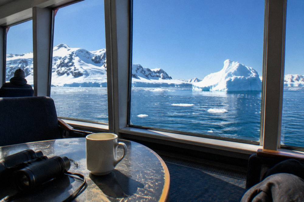A bright expedition ship lounge with large windows looking out over icebergs