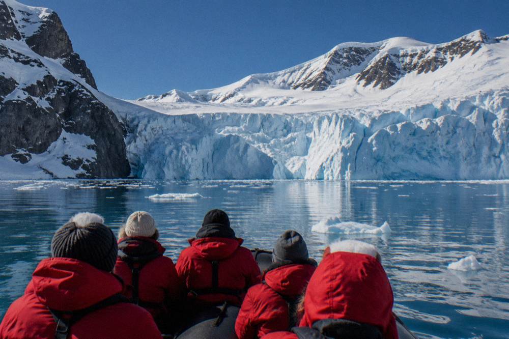 A Zodiac full of red-jacketed passengers approaching a glacier face in a calm Antarctic bay