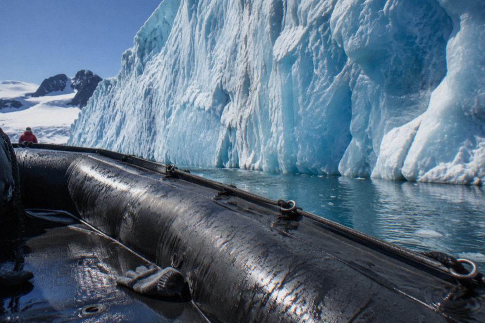 A Zodiac cruising past a wall of blue glacier ice in a calm Antarctic bay