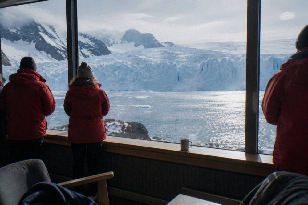 Passengers in expedition parkas watching icebergs from a ship's observation lounge windows
