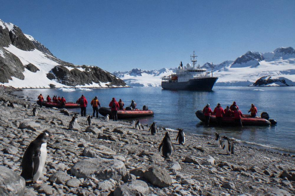 A small expedition ship with red Zodiac boats clustered along the shoreline of a rocky Antarctic bay