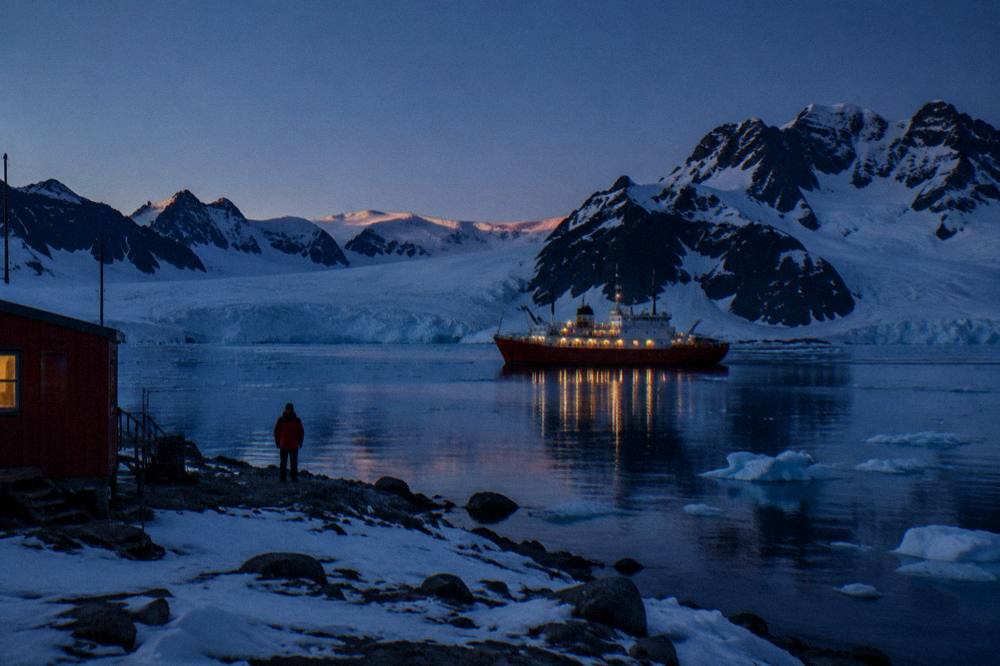 A small red-hulled expedition ship at anchor in a bay surrounded by sea ice and mountains